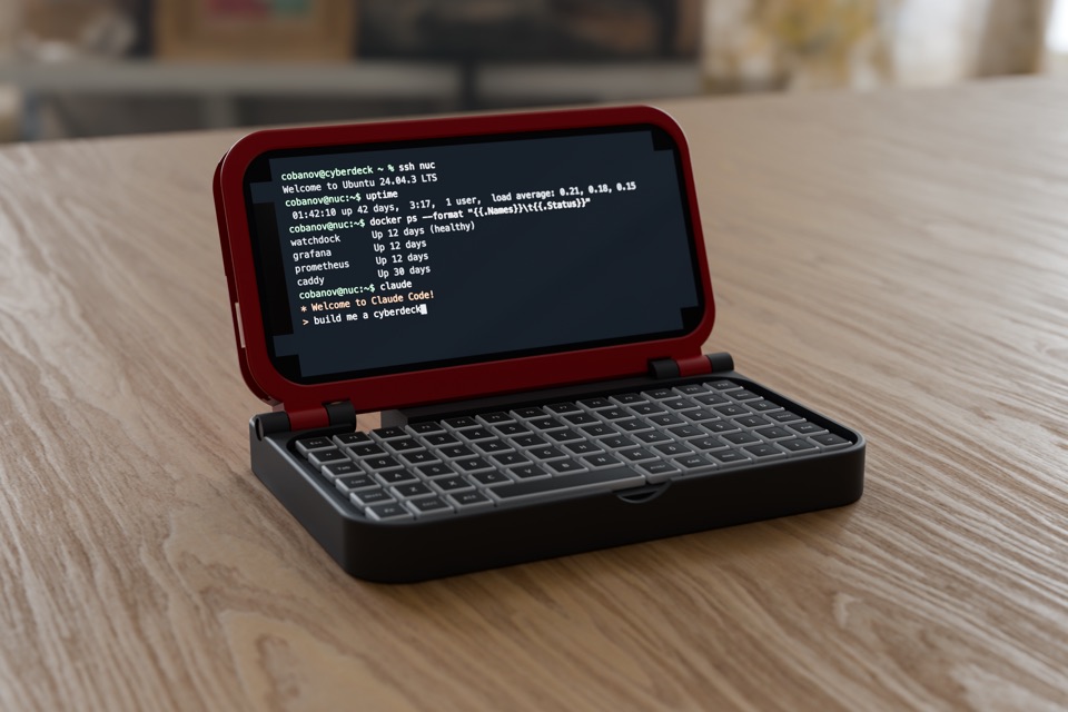 A red and black 3D-printed clamshell deck, open on a wooden table: an iPhone in the lid shows a terminal, a compact keyboard sits in the base.